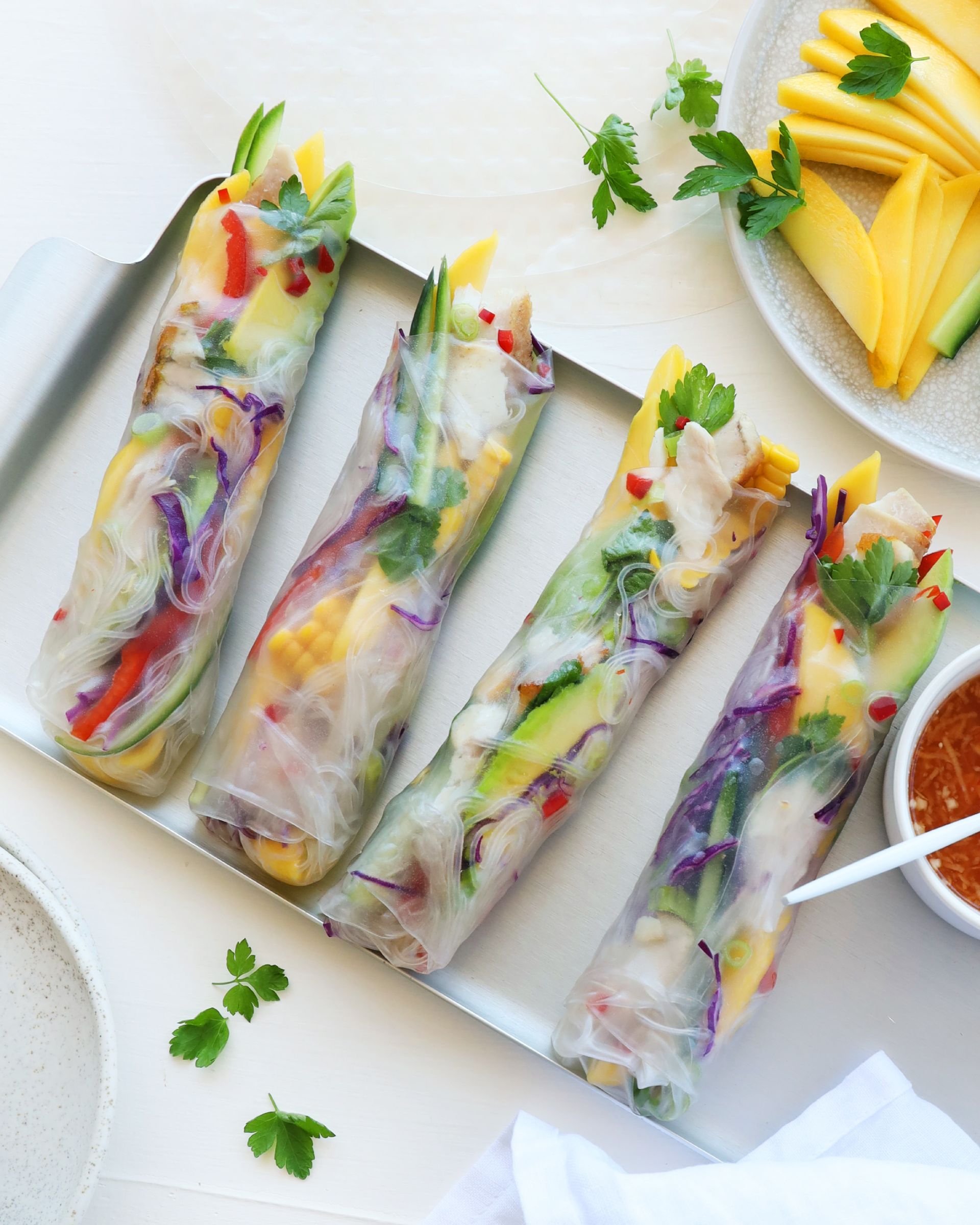Four fresh spring rolls filled with colorful vegetables and mango, served on a metal tray with dipping sauce.