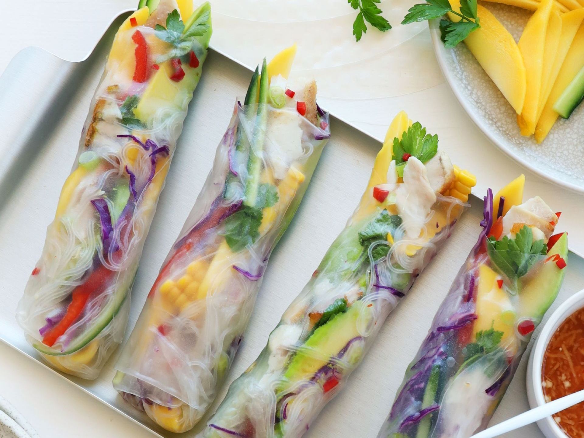 Four fresh spring rolls filled with colorful vegetables, mango, and chicken on a tray next to a side of dipping sauce.