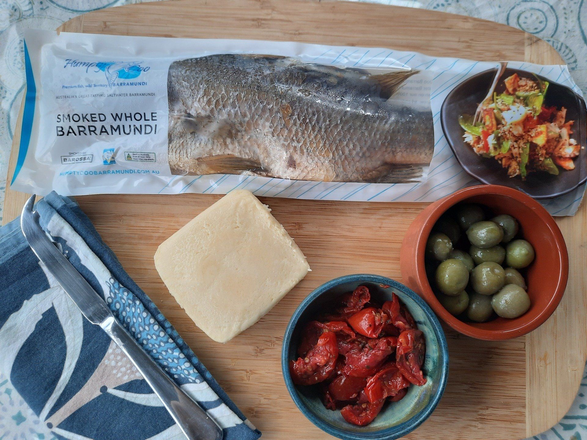 A wooden cutting board with a packaged whole barramundi, a block of cheese, green olives, sun-dried tomatoes, and salad.