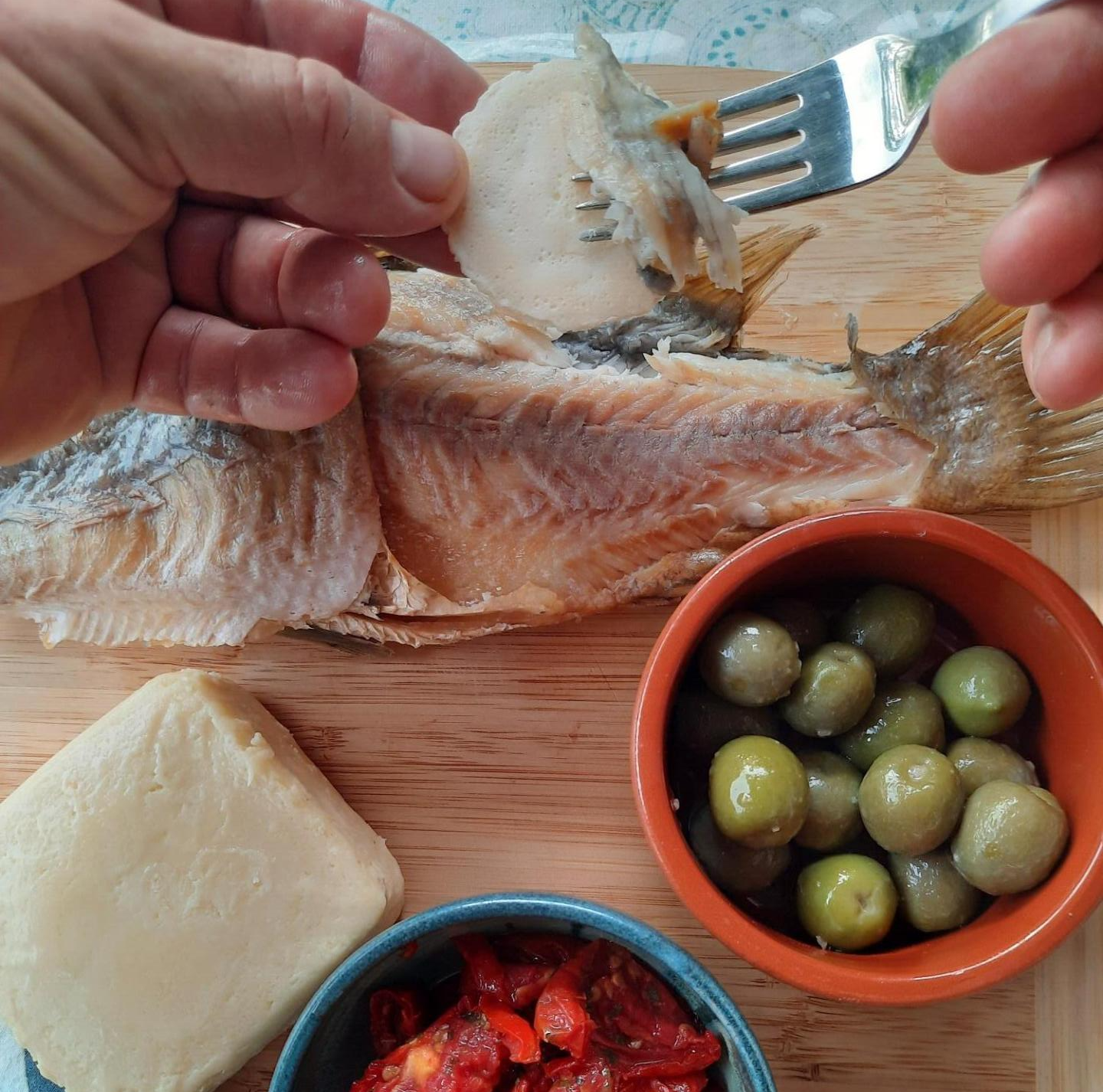 Hands use a fork to flake a cooked white fish fillet on a wood board next to a block of cheese, olives, and red peppers.