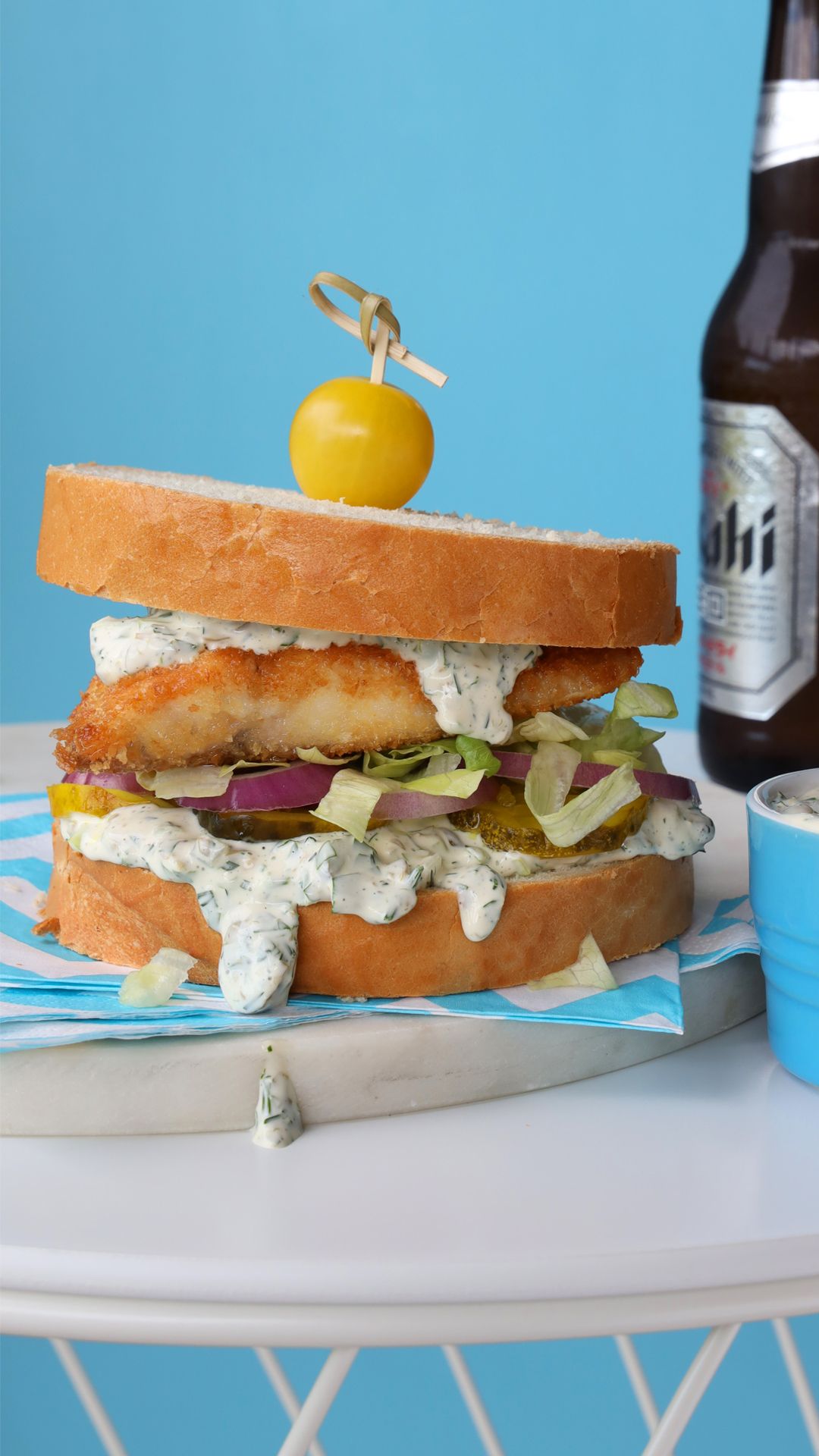 A fried fish sandwich topped with a yellow cherry tomato, sitting on a blue patterned napkin next to a bottle of beer.