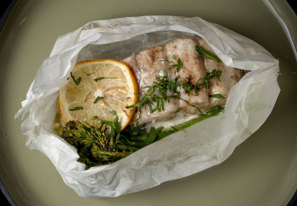 A fillet of white fish served in parchment paper with a lemon slice, fresh herbs, and green vegetables on a green plate.