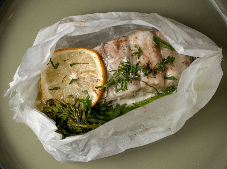 A piece of white fish with a lemon slice and greens cooked in a parchment paper packet, served on a green plate.
