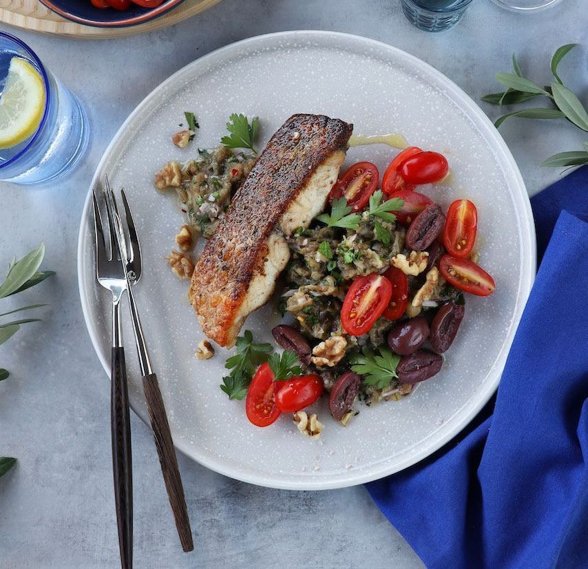 A seared fish fillet served on a bed of herbs, walnuts, olives, and halved cherry tomatoes on a light-colored plate.