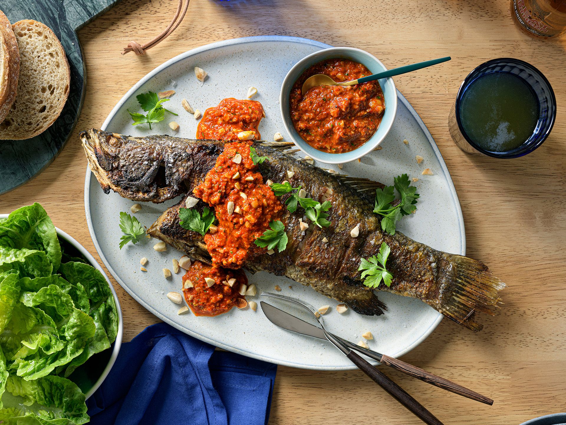 A grilled whole fish topped with red sauce and herbs on a plate, served with lettuce and bread on a wooden table.