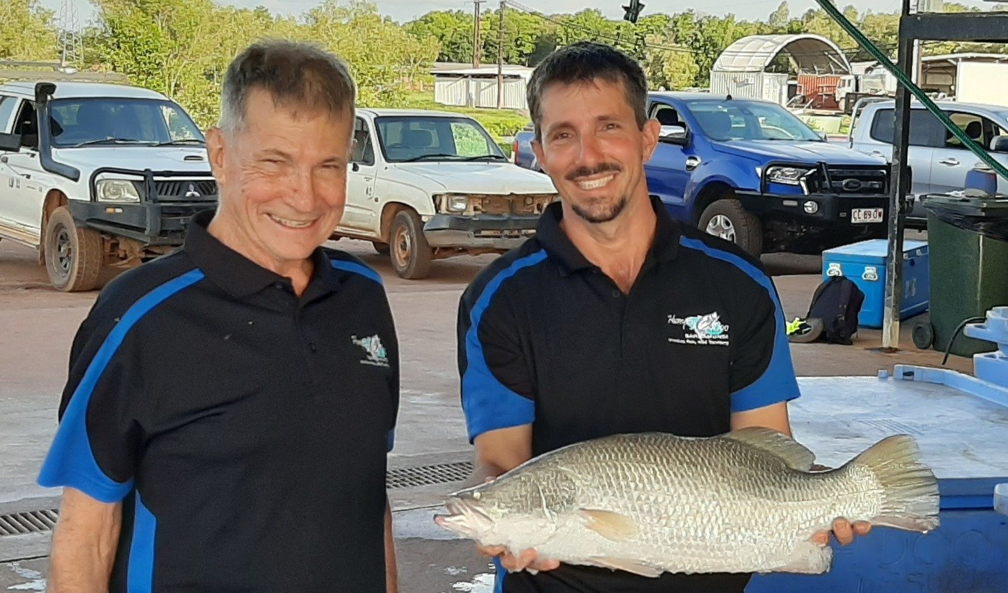 Two people in matching blue and black polo shirts hold a large fish in front of a parking lot with vehicles.