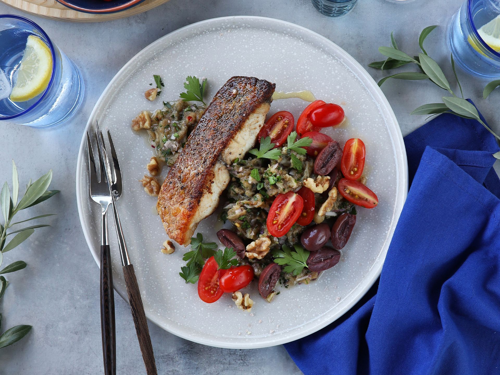 A plate of seared fish served with lentils, walnuts, cherry tomatoes, and herbs, set on a grey table with a blue napkin.