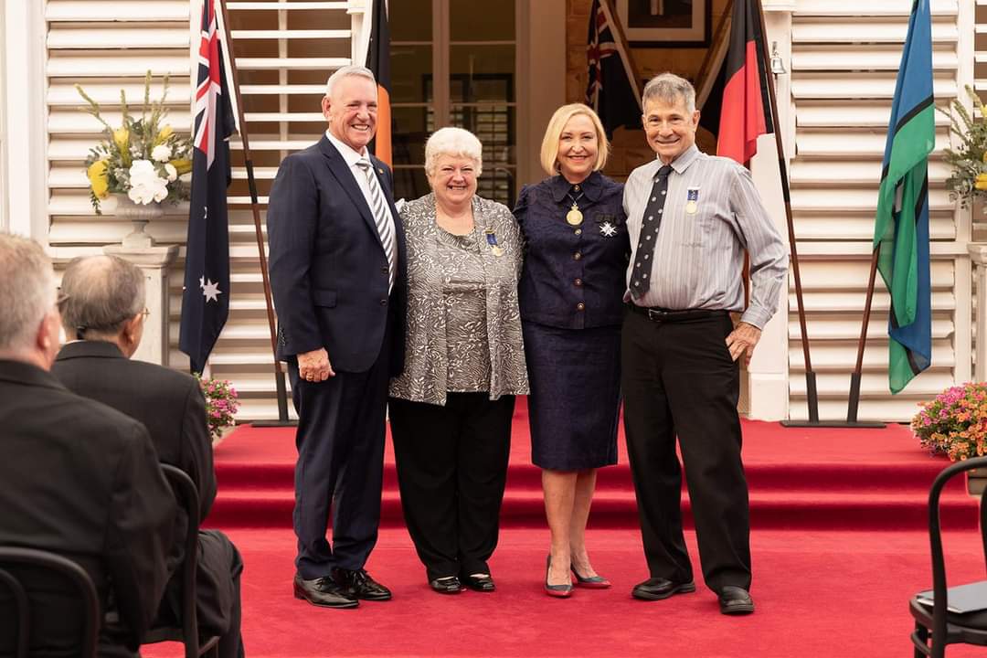 Four people stand together on a red carpeted stage in front of flags, posing for a group portrait at an awards ceremony.