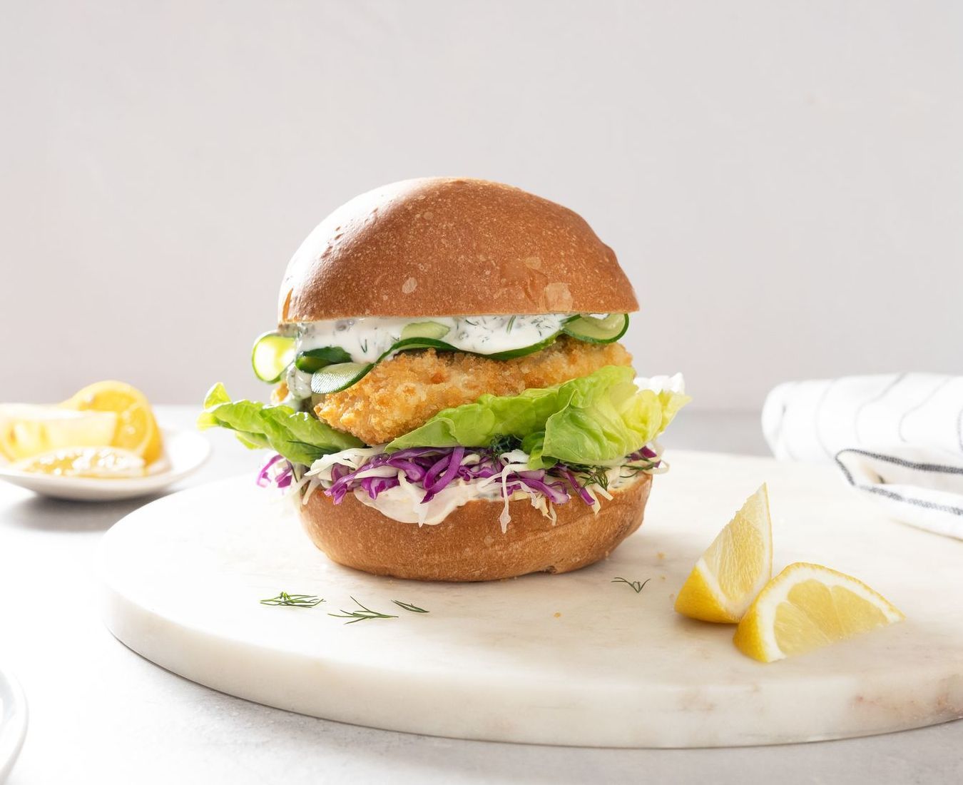 A breaded fish sandwich on a round bun with lettuce, red cabbage, and cucumber slices, served with lemon wedges.