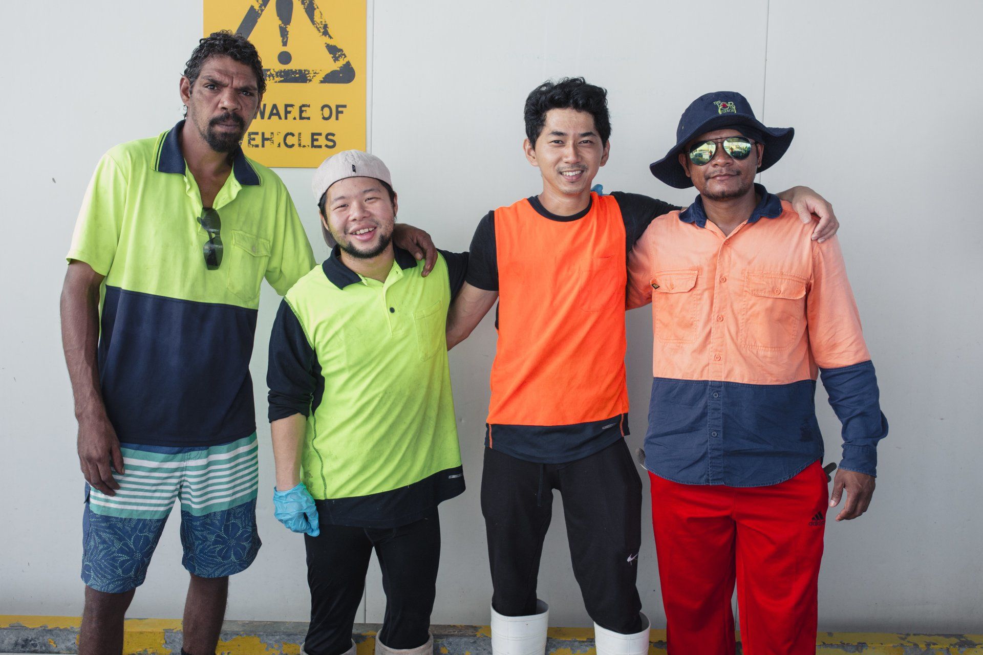 Four people in high-visibility work uniforms stand side-by-side against a wall with a warning sign.