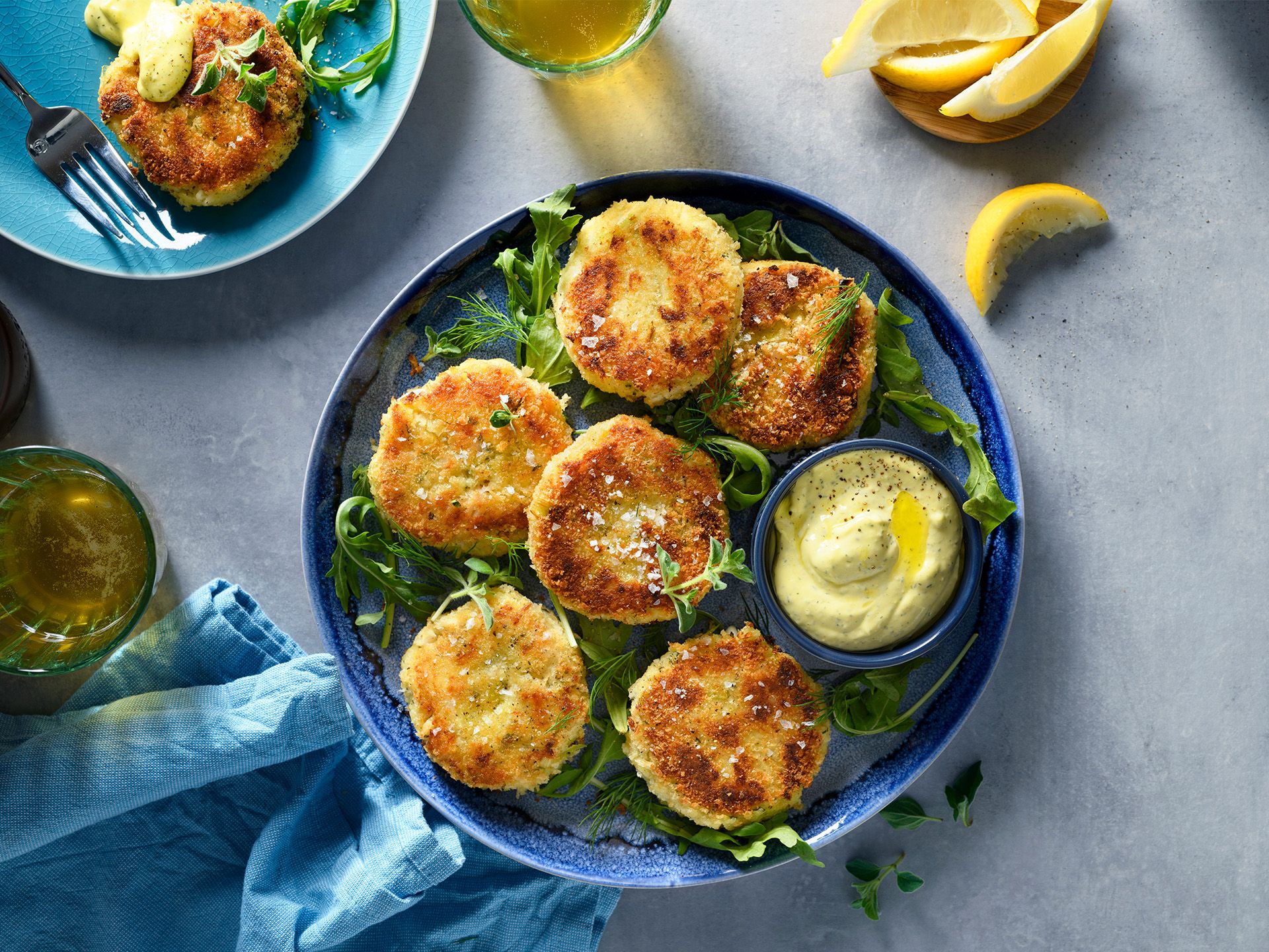 A blue plate of golden-brown fried crab cakes on a bed of greens, served with a small bowl of creamy yellow dipping sauce.