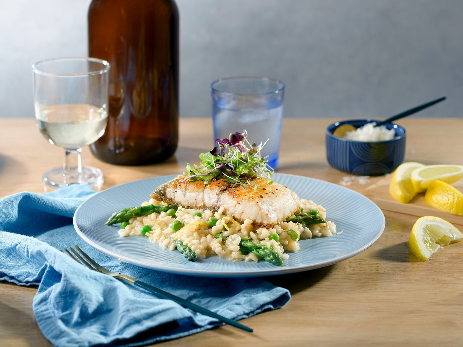 A plate of risotto topped with a seared fish fillet and greens, served with a drink and lemon wedges on a wooden table.