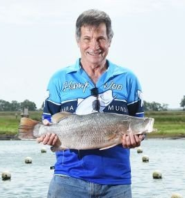 A smiling angler in a blue fishing shirt holds a large barramundi fish in front of a body of water.