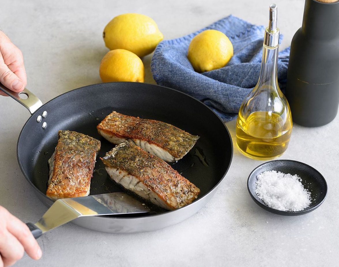 A person uses a spatula to flip seasoned fish fillets in a frying pan, with lemons, oil, and sea salt nearby.