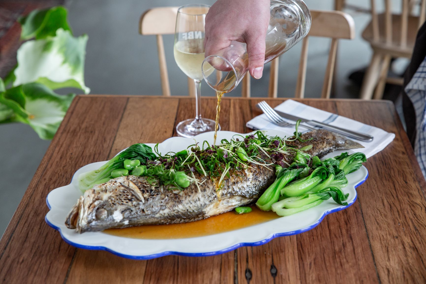 A hand pours sauce over a whole roasted fish served with bok choy on a wooden table next to a glass of white wine.