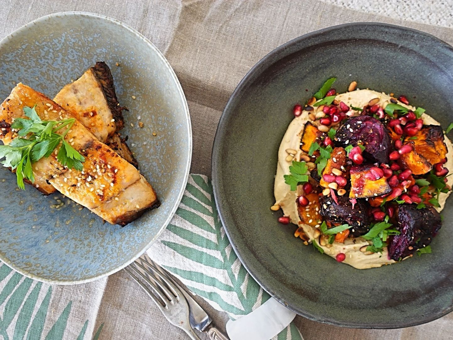 Two bowls on a linen cloth: one holds seared fish garnished with herbs, the other features roasted beets over hummus.