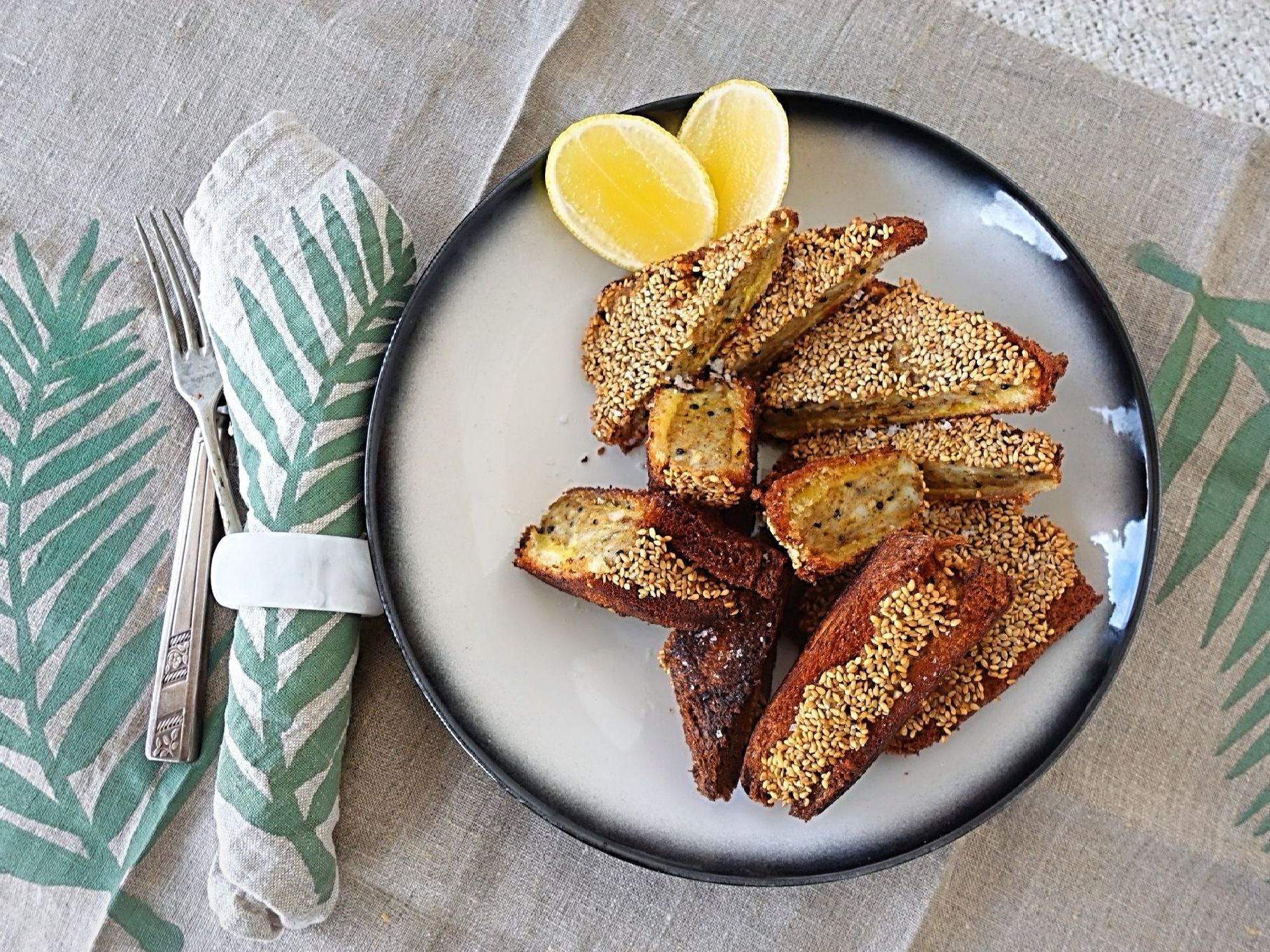 A plate of sesame-crusted fried food triangles with two lemon wedges, next to a patterned napkin and fork on a table.