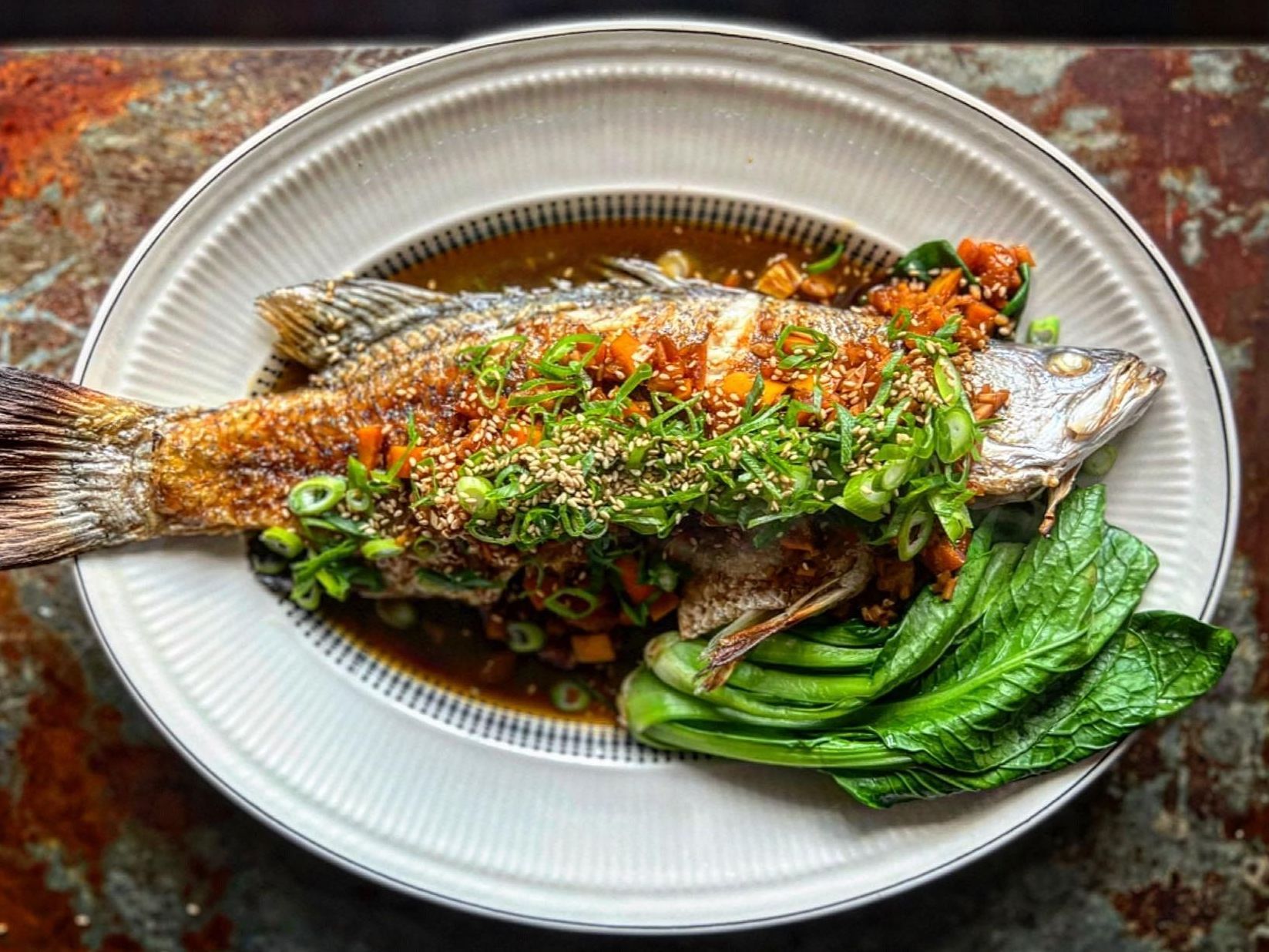 A whole fried fish served on a platter with a savory sauce, garnished with scallions, and accompanied by bok choy.