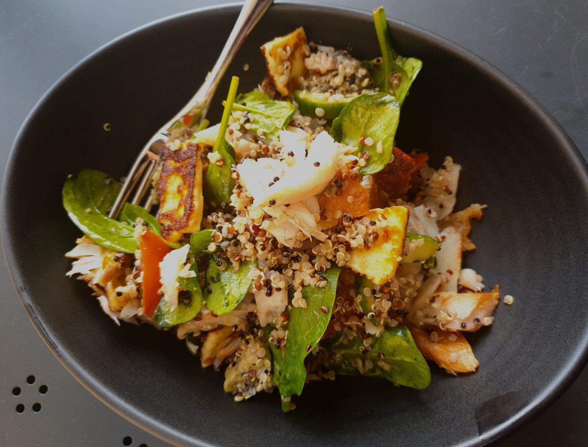 A bowl of quinoa salad with spinach, seared halloumi cheese, and pieces of flaked fish, served with a fork.