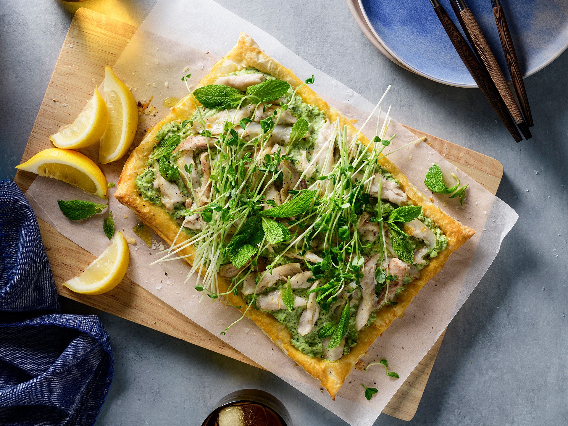 A rectangular puff pastry tart topped with chicken, green herbs, and sprouts, served on a wooden board with lemon wedges.