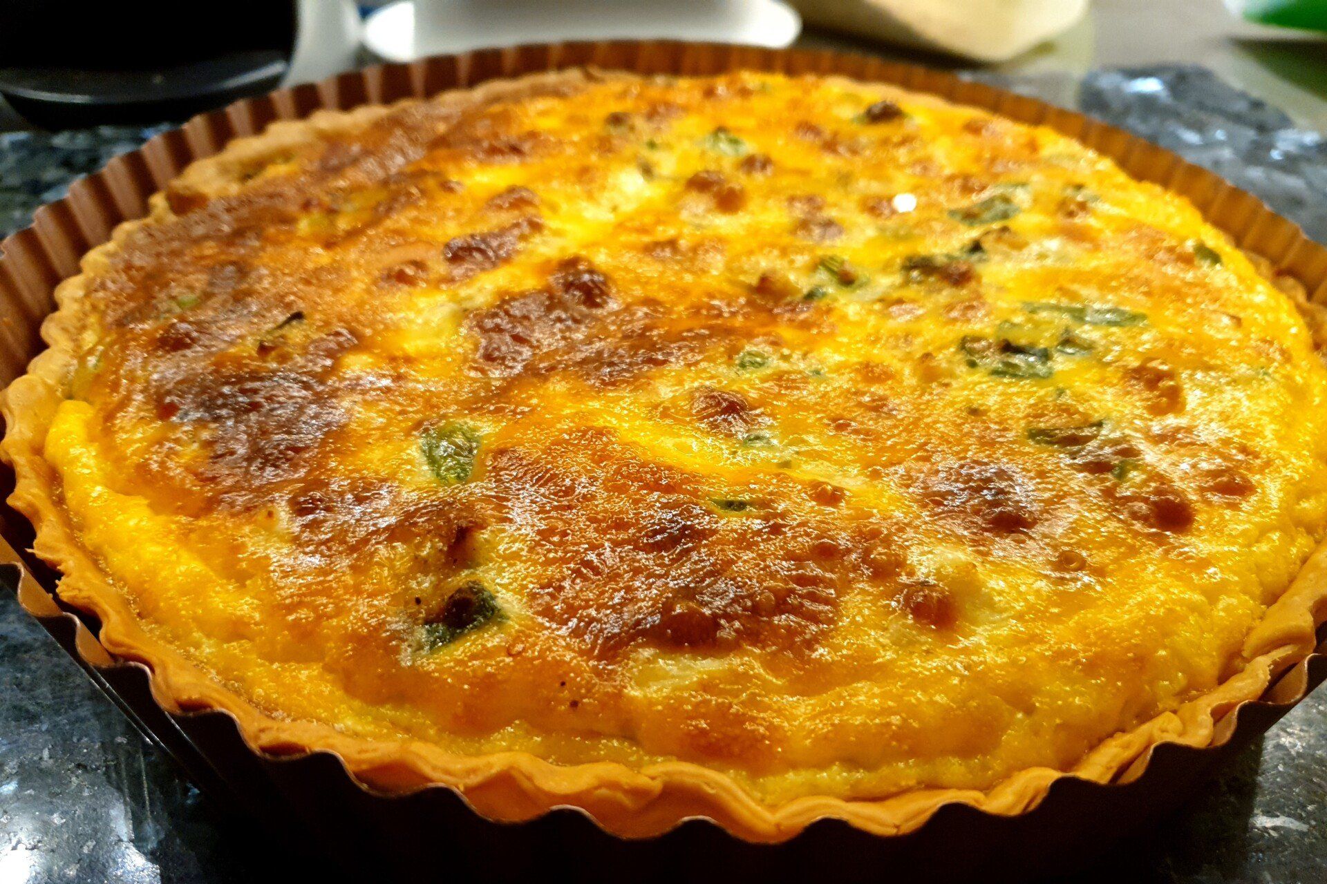 A golden-brown baked quiche with green flecks, served in a fluted tart pan on a dark, textured surface.