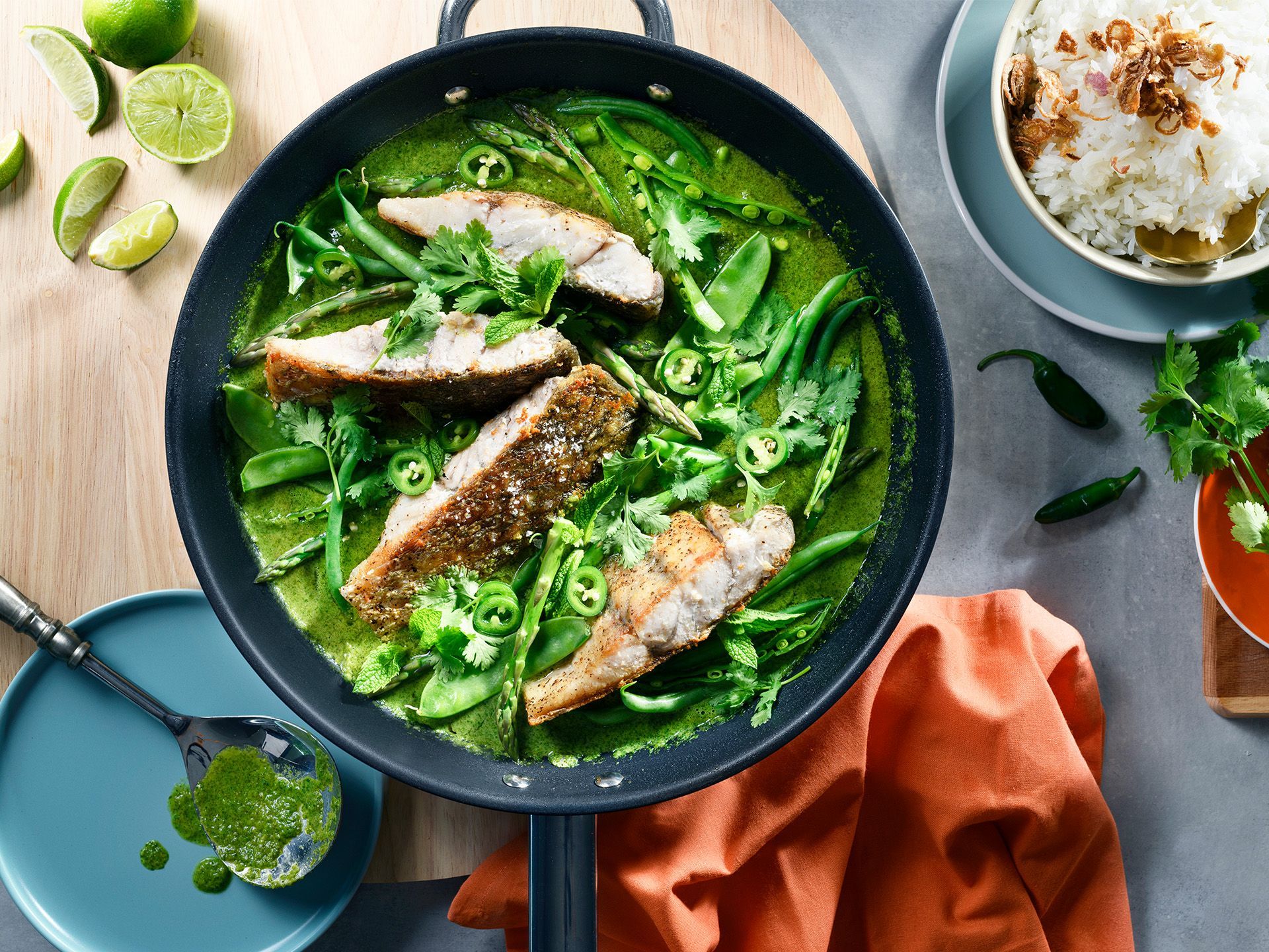 A pan of green Thai curry with seared fish, lime wedges, and a bowl of rice on a tabletop.