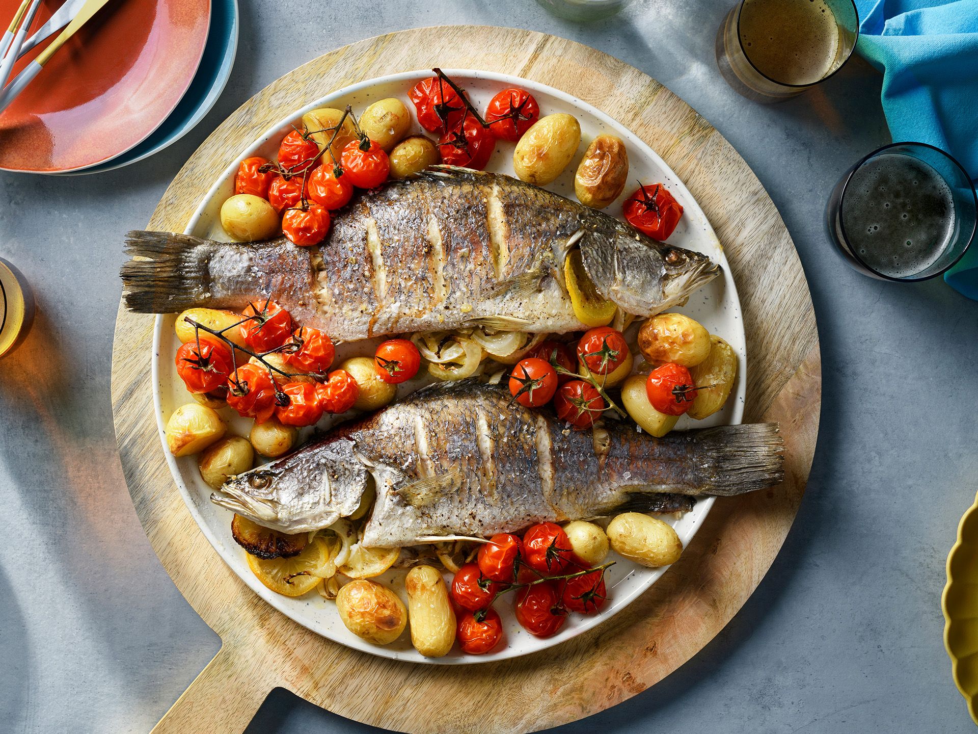 Two whole roasted fish served on a white plate with cherry tomatoes and small potatoes, set on a wooden cutting board.