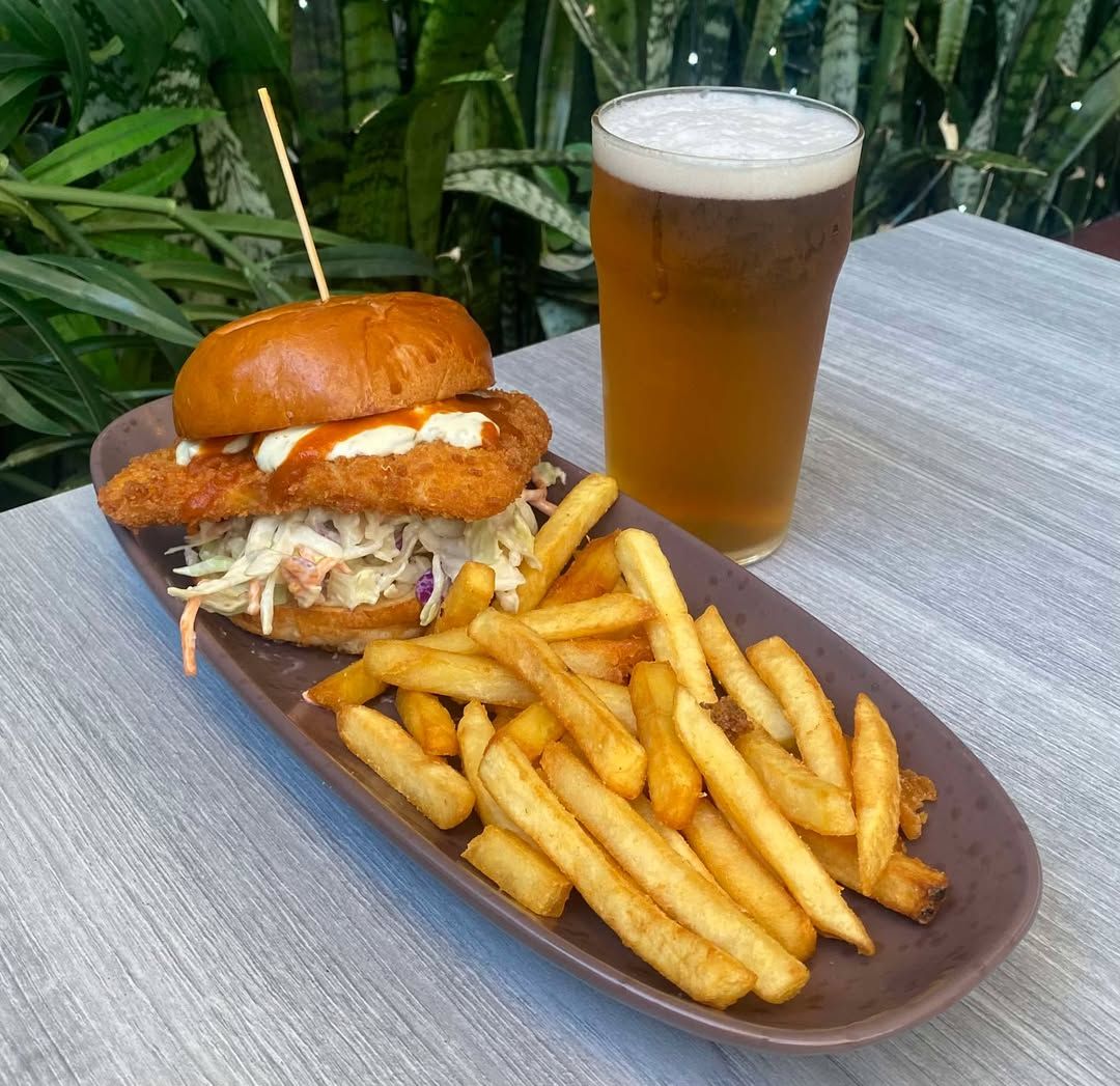 A breaded chicken sandwich with coleslaw and a side of fries on a long plate next to a glass of beer.