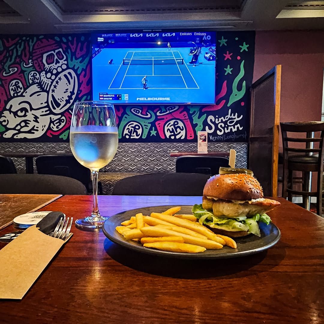 A burger and fries with a glass of white wine on a wooden table, in front of a wall with colorful graffiti and a TV.