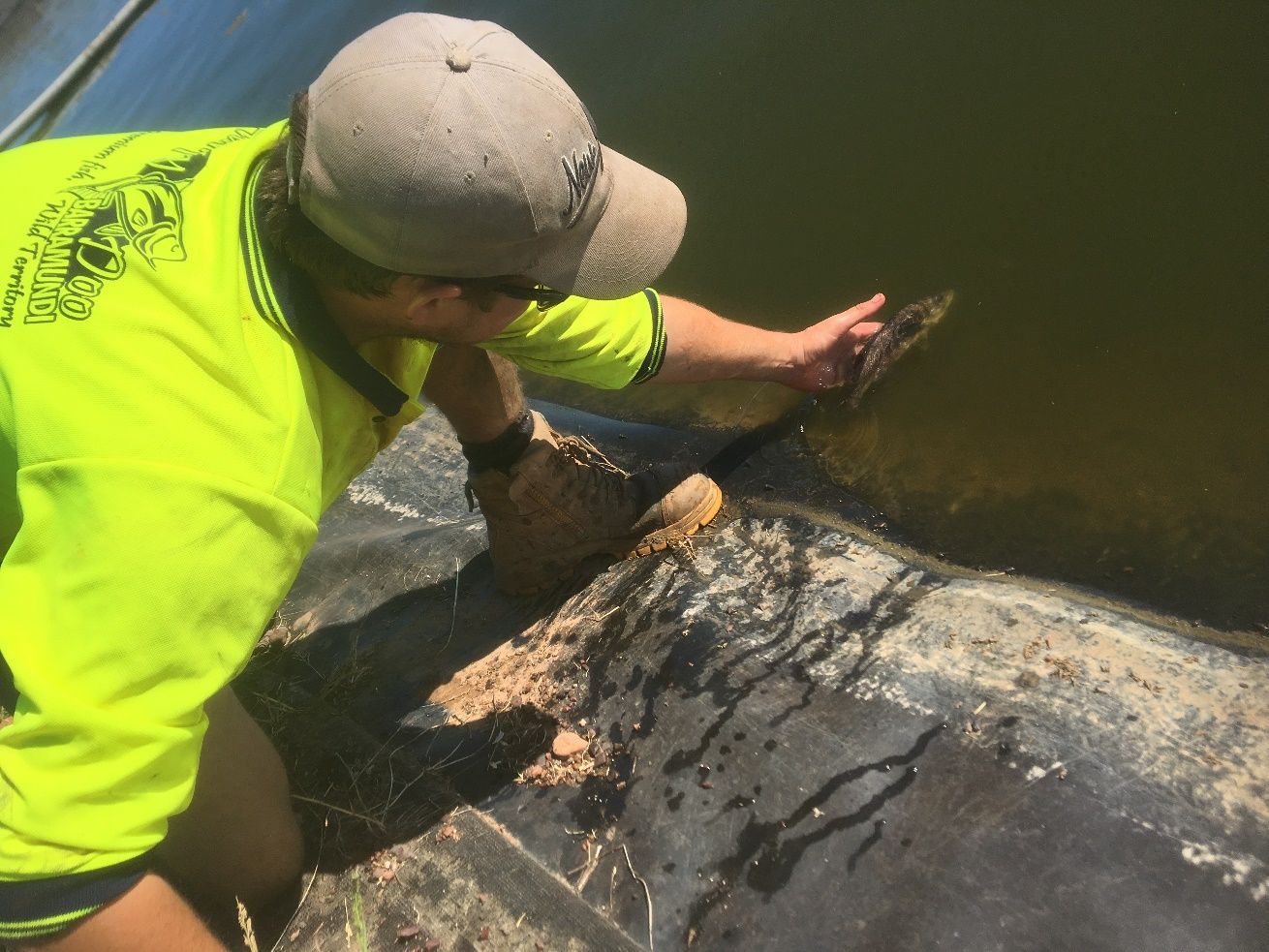 A person wearing a bright yellow shirt and cap kneels at a water's edge, using a tool to reach into the dark water.
