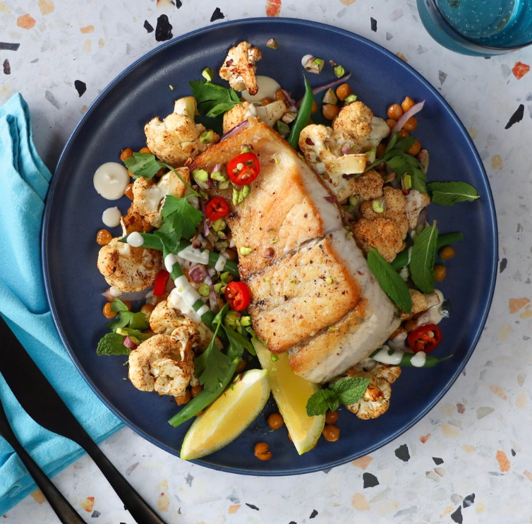 Pan-seared fish fillet served with roasted cauliflower, chickpeas, fresh herbs, chili slices, and lemon wedges on a plate.
