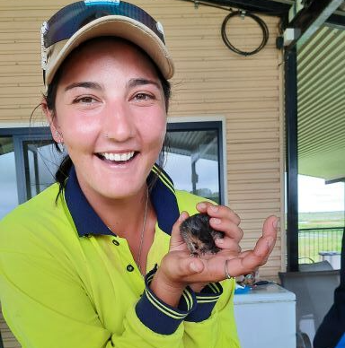 A smiling person in a high-visibility lime shirt and cap holds a small, dark animal in their cupped hands outdoors.