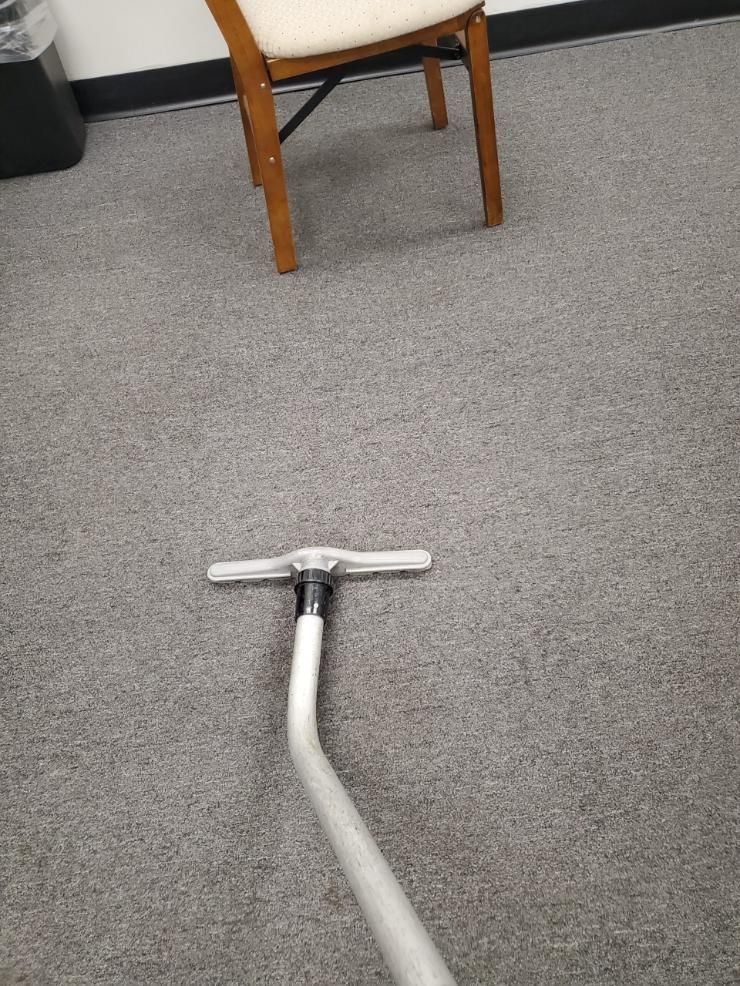Silver vacuum cleaner nozzle on gray carpet beneath a wooden chair.