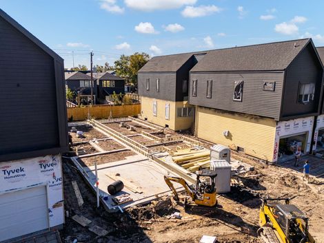 Construction site with two houses, exposed foundation, debris, and a yellow excavator under a blue sky
