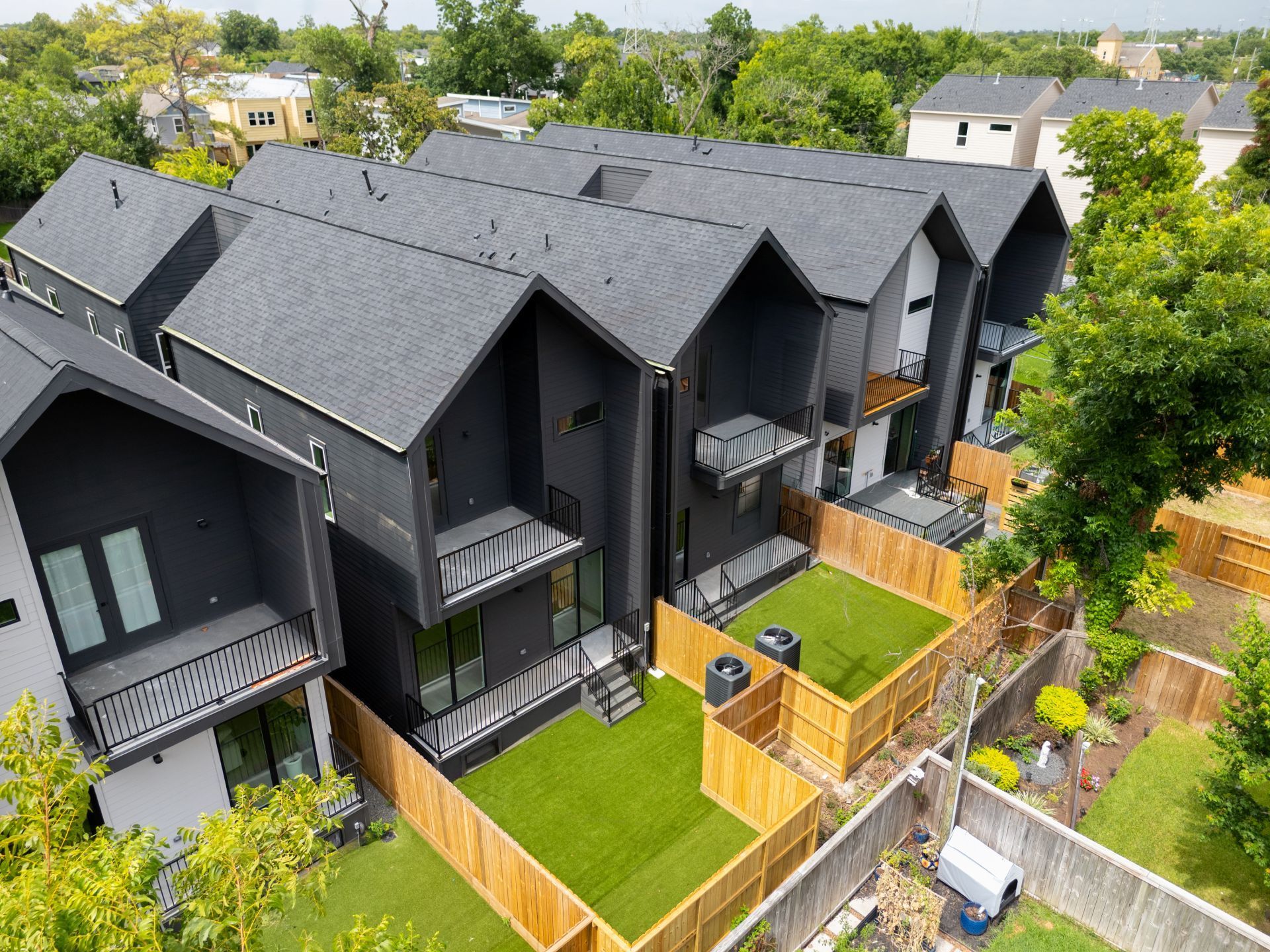 Aerial view of modern dark-gray townhouses with balconies, fenced yards, and green lawns.