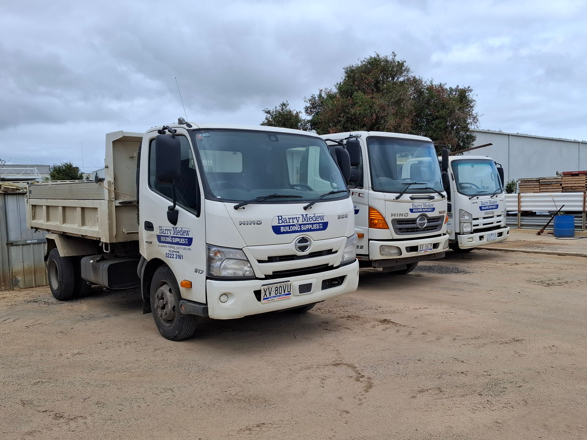 Three white dump trucks parked on a gravel lot under a cloudy sky.