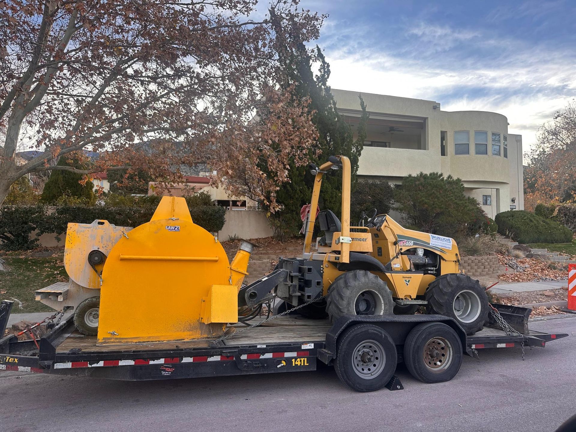 Yellow construction equipment on a flatbed trailer in front of a building and trees