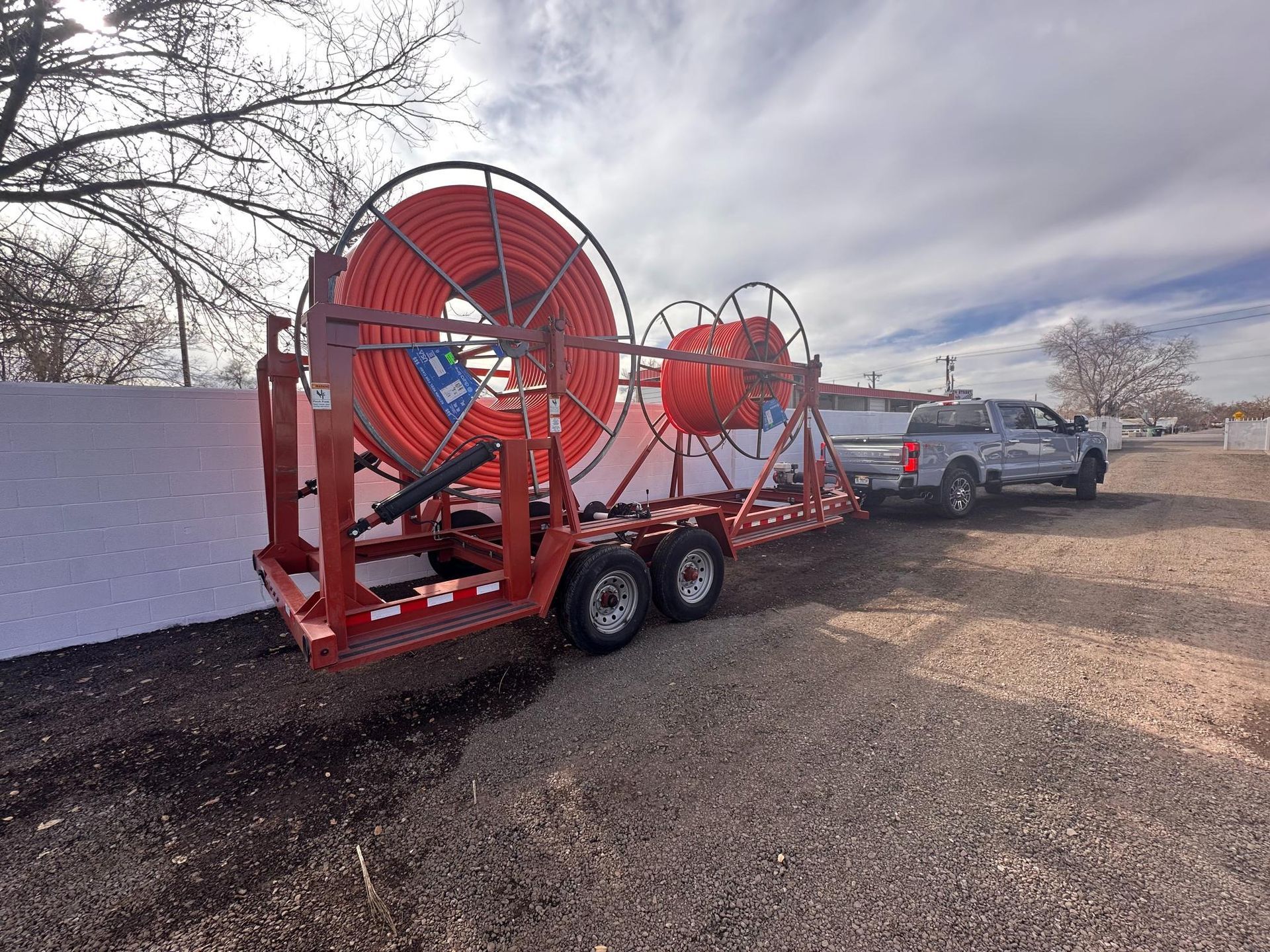 Red industrial cable reel trailer parked on gravel beside a white wall, with a pickup truck in the background.