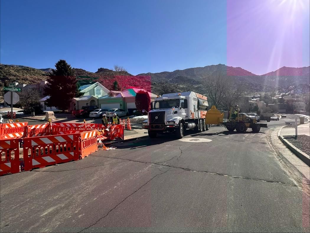 Road construction site with orange barricades, a truck, and workers on a sunny street with hills in the background