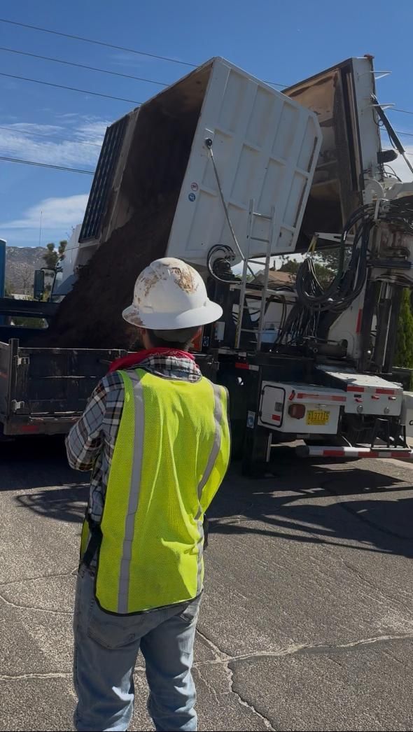 Worker in hard hat and neon vest watching a truck unload a large metal structure outdoors.