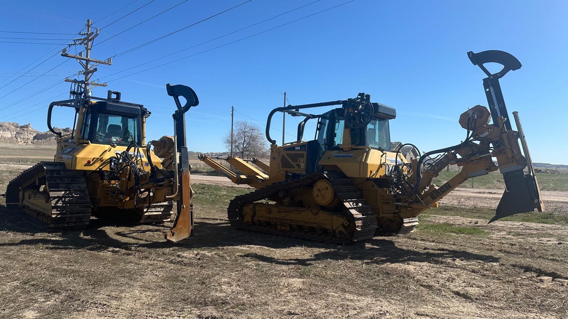 Two yellow bulldozers parked on a dirt lot under a clear blue sky, one with a raised rear attachment.