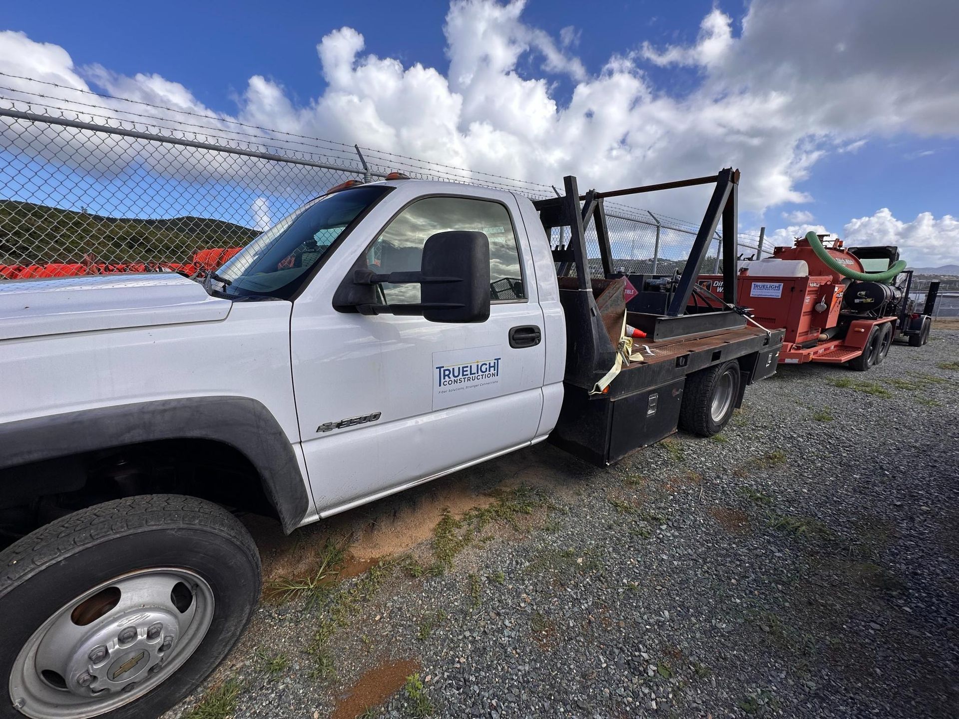 White flatbed truck parked on gravel beside red farm equipment under a cloudy sky