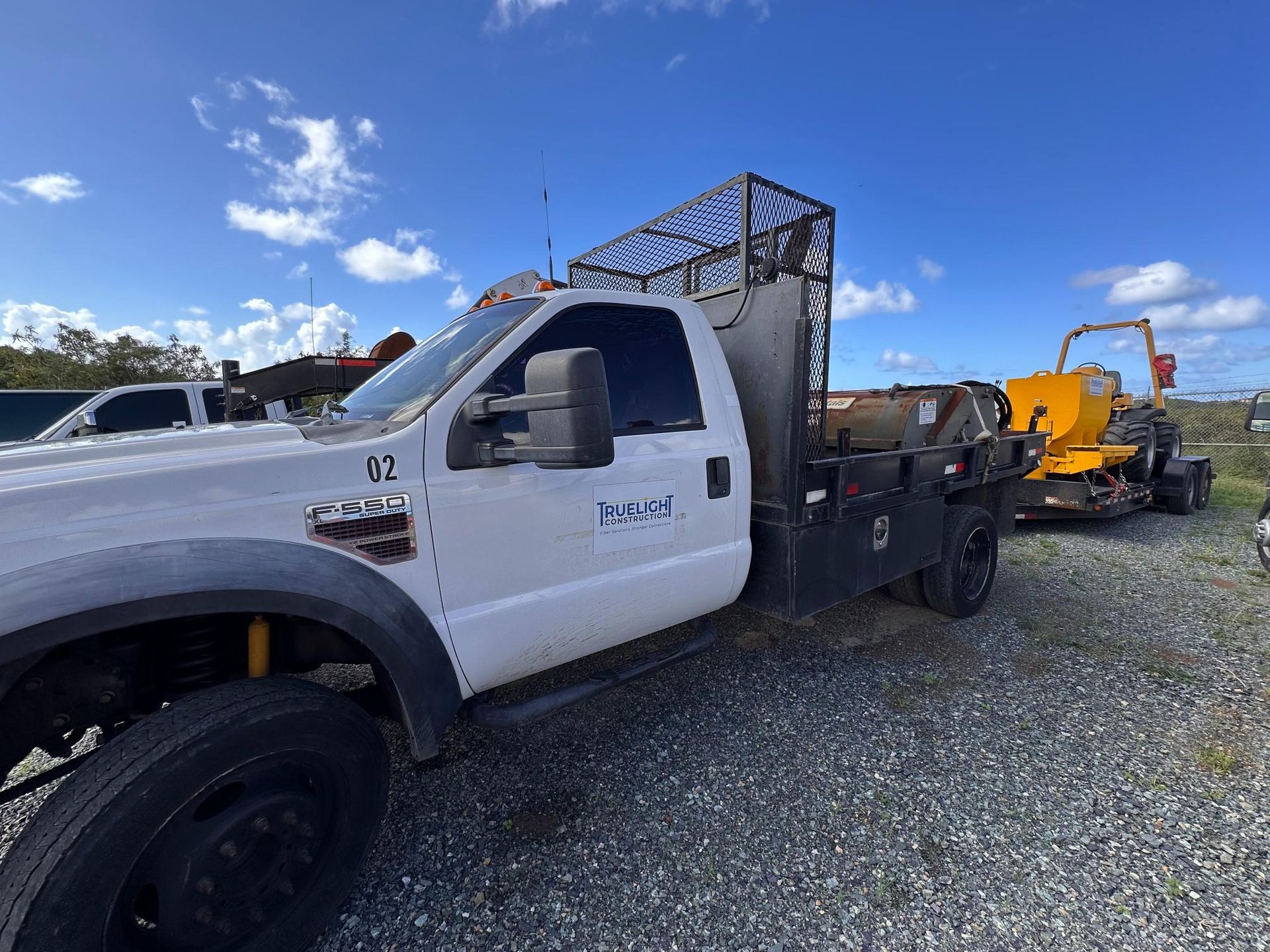 White work truck with metal rack parked on gravel beside heavy equipment under a blue sky