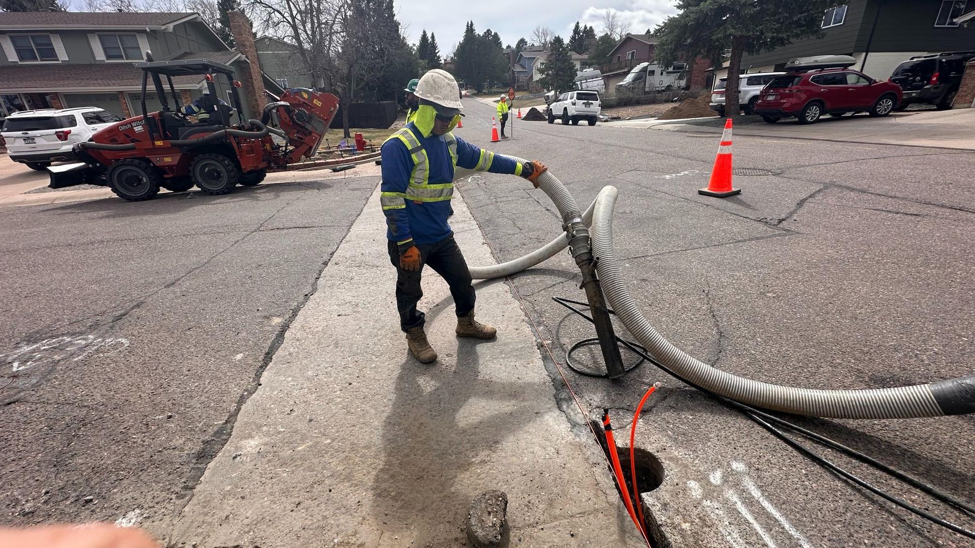 Worker in safety gear sprays water beside a large hose on a street near parked cars and cones.