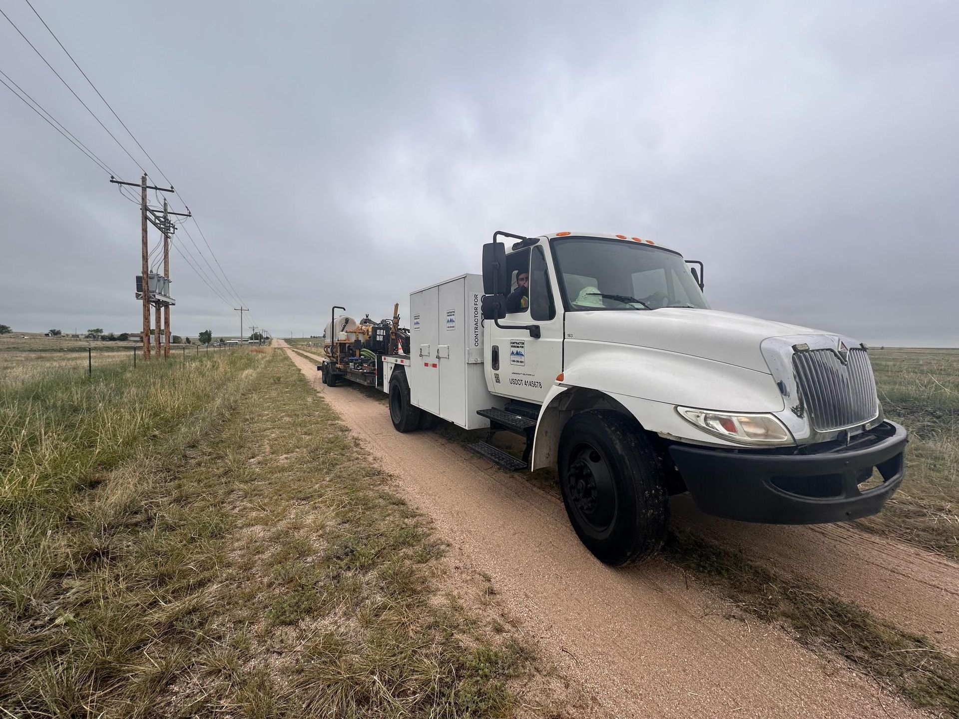 White tow truck with trailer parked on a gravel road beside grassy fields under a cloudy sky