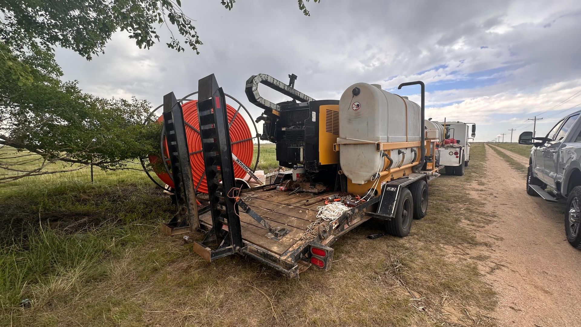 Heavy farm sprayer trailer parked by a roadside, with red reel, hoses, and white tank under cloudy sky
