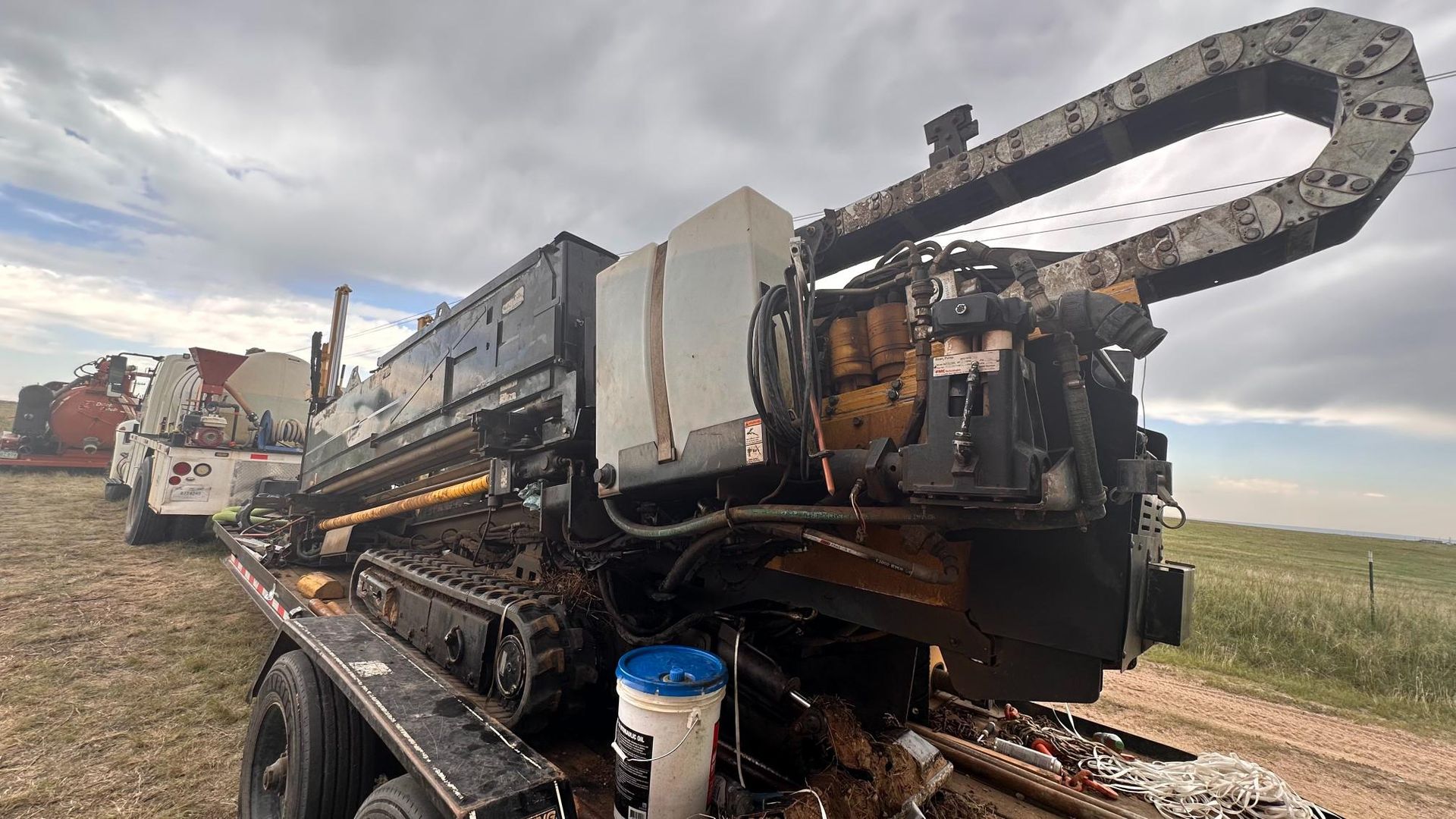 Destroyed drilling machine on a muddy roadside, with smoke or steam rising under a cloudy sky