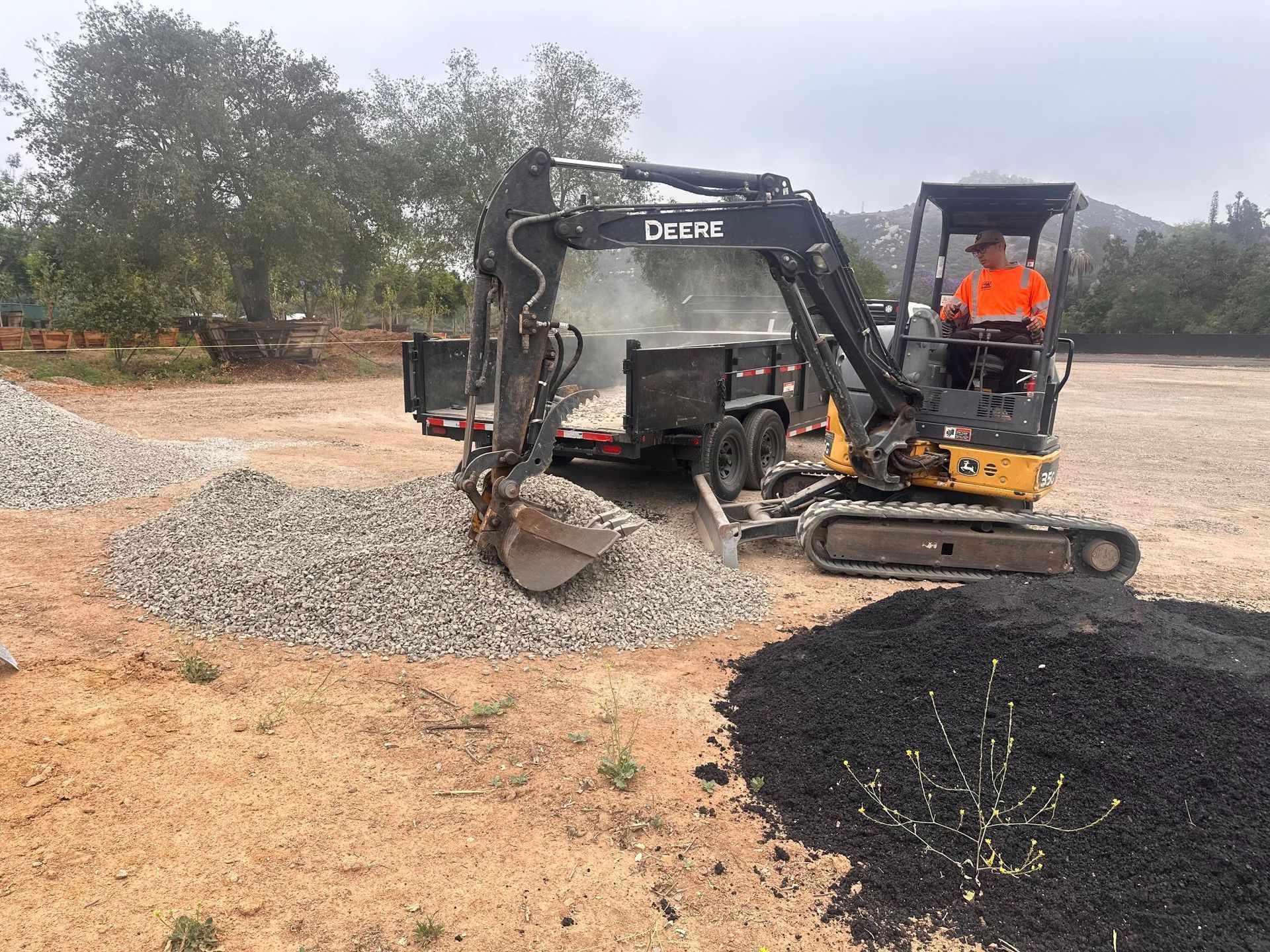 Mini excavator spreading gravel on a dirt lot, with an operator in orange safety gear.