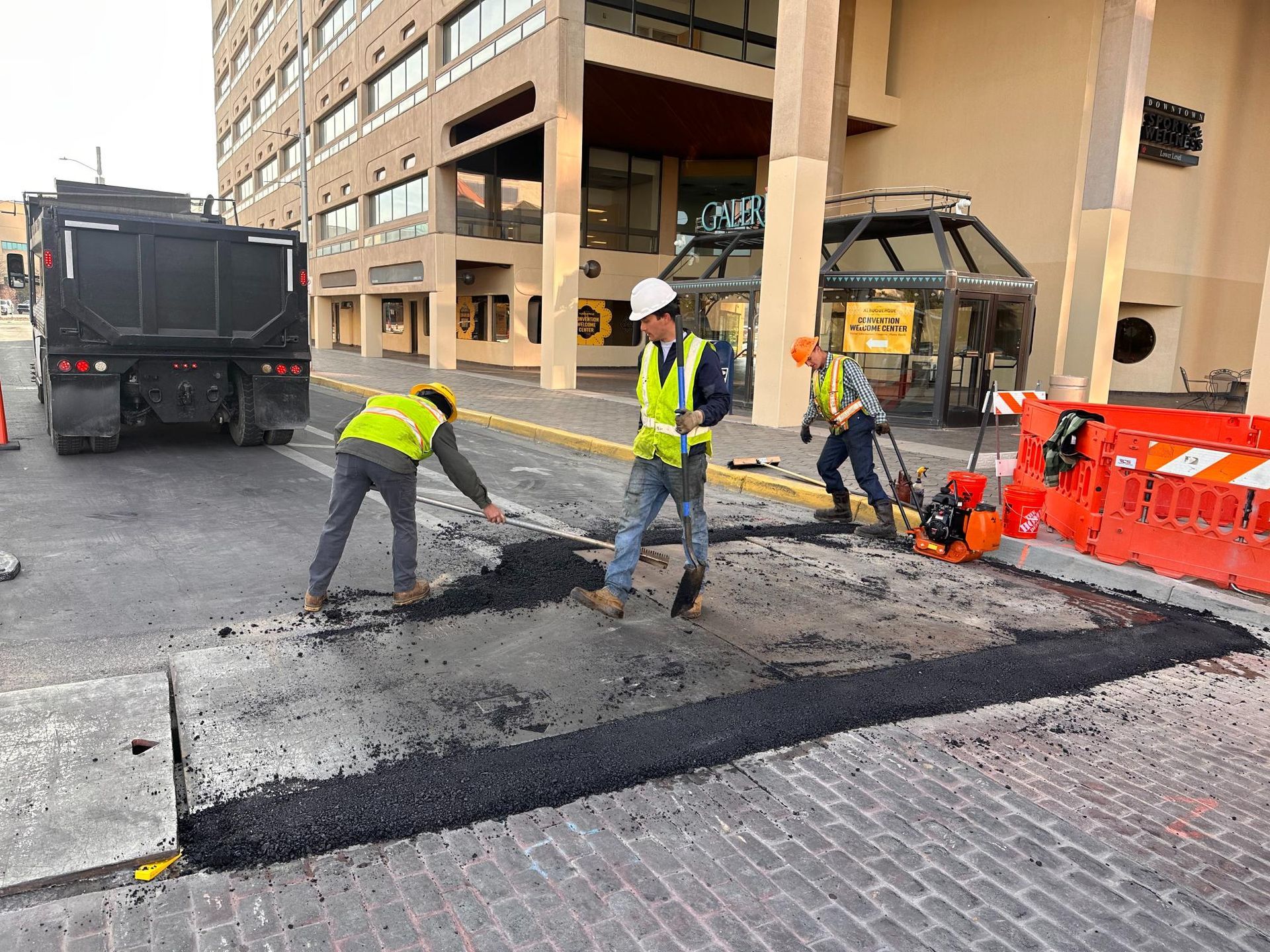 Road repair crew paving a damaged street beside a building, with orange barriers and trucks nearby