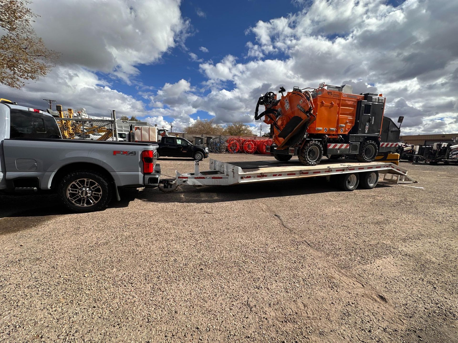 Silver pickup towing a flatbed trailer with orange heavy machinery in a gravel lot under cloudy skies